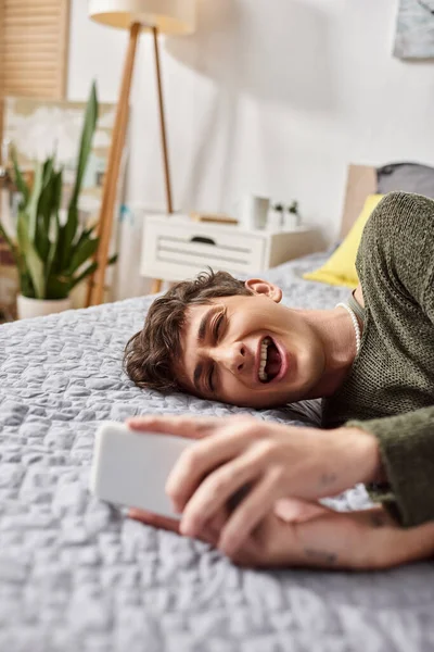 Blogueiro transgênero animado com cabelo encaracolado usando smartphone, deitado na cama, influenciador de mídia social — Fotografia de Stock