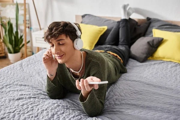 Feliz encaracolado transgênero pessoa em fones de ouvido sem fio segurando smartphone no quarto, ouvir música — Fotografia de Stock