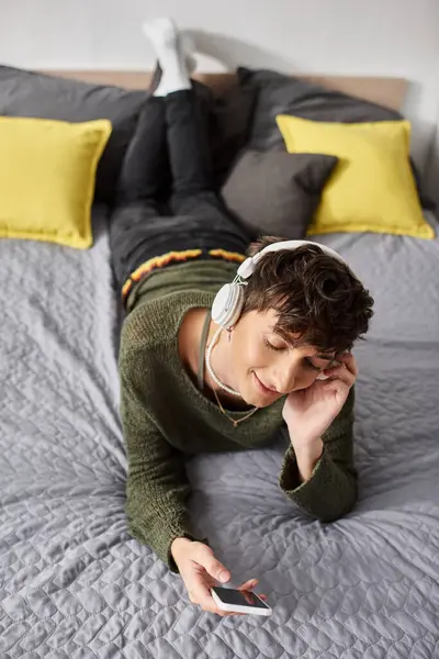 Happy curly transgender person in wireless headphones holding smartphone in bedroom, listen music — Stock Photo