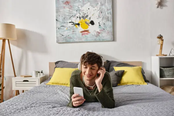 Happy curly transgender person in wireless headphones holding smartphone in bedroom, listen music — Stock Photo