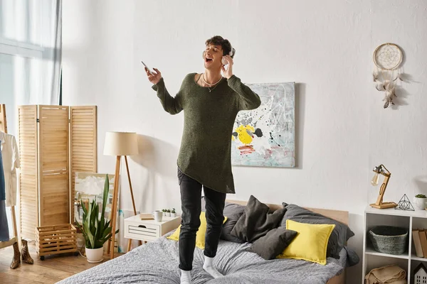 Happy curly transgender person in wireless headphones holding smartphone and standing on bed — Stock Photo
