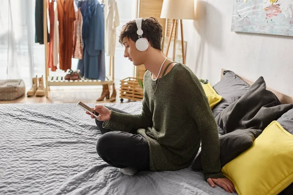 Encaracolado transgênero pessoa em fones de ouvido sem fio usando smartphone e sentado na cama, mídias sociais — Fotografia de Stock