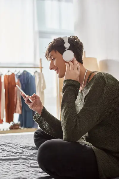 Happy curly transgender person in wireless headphones holding smartphone and sitting on bed — Stock Photo