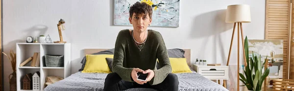 Banner di giovane transgender in abbigliamento casual guardando la fotocamera e seduto sul letto a casa — Foto stock