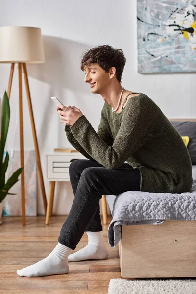 Cheerful and curly transgender influencer using smartphone and sitting on bed, social media — Stock Photo