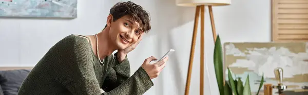 Banner of happy curly transgender influencer using smartphone and sitting in bedroom, social media — Stock Photo