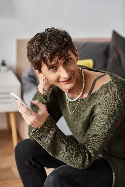 Happy and curly transgender influencer using smartphone and sitting on bed, social media — Stock Photo