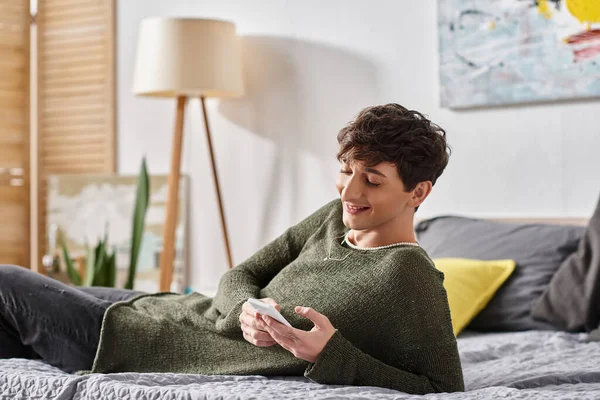 Cheerful and curly transgender blogger using smartphone and lying on bed, social media influencer — Stock Photo
