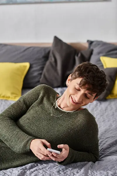 Cheerful and curly transgender blogger using smartphone and lying on bed, social media influencer — Stock Photo