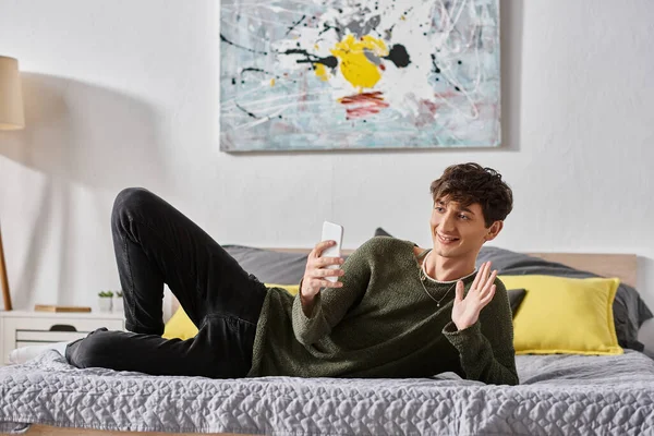 Happy and curly transgender social media influencer waving hand during online streaming in bedroom — Stock Photo