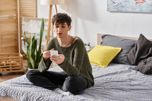 Happy and curly transgender influencer using smartphone and sitting on bed, social media — Stock Photo