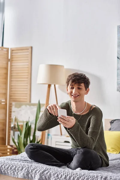 Happy and curly transgender influencer using smartphone and sitting on bed, social media — Stock Photo