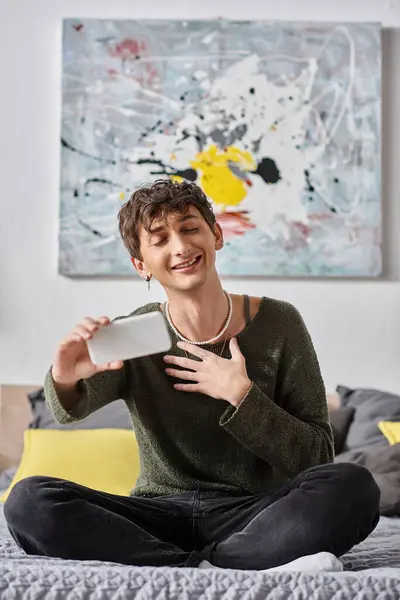 Happy curly transgender influencer smiling and using smartphone while sitting on bed, social media — Stock Photo