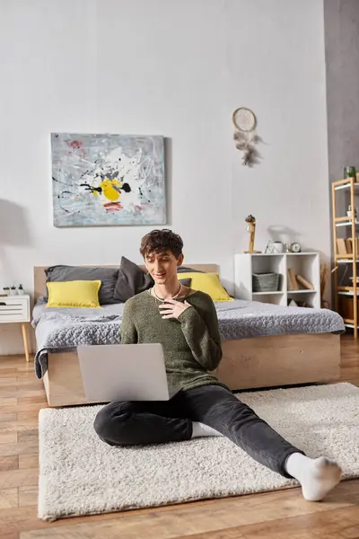 Feliz y rizado transgénero influencer utilizando el ordenador portátil y sentado en la alfombra cerca de la cama, blogger - foto de stock