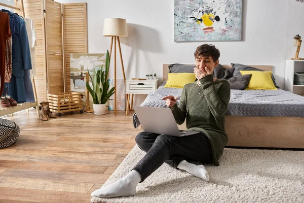 Joyful curly transgender influencer covering mouth and using laptop while sitting near bed, blogger — Stock Photo