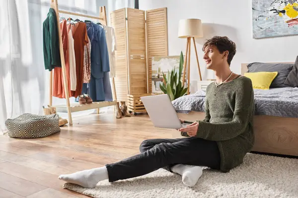 Happy and curly transgender influencer using laptop and sitting on carpet near bed, blogger — Stock Photo