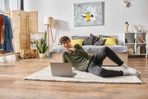 Happy and curly transgender influencer using laptop and lying on carpet near bed, social media — Stock Photo