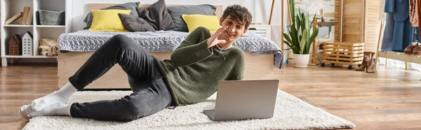 Happy and curly transgender influencer using laptop and sitting on carpet near bed, blogger banner — Stock Photo