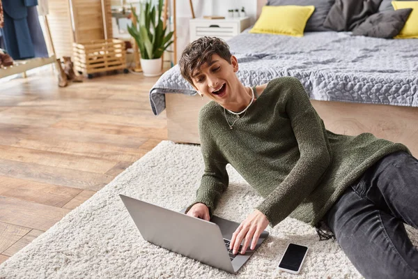 Happy curly transgender influencer using laptop and sitting on carpet next to smartphone, blogger — Stock Photo