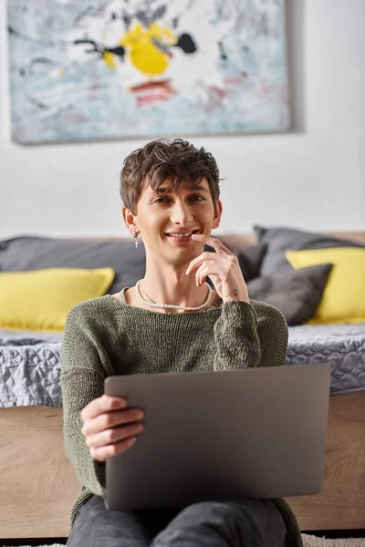 Zufriedene und lockige Transgender-Influencerin mit Laptop und neben dem Bett sitzend, soziale Medien — Stockfoto