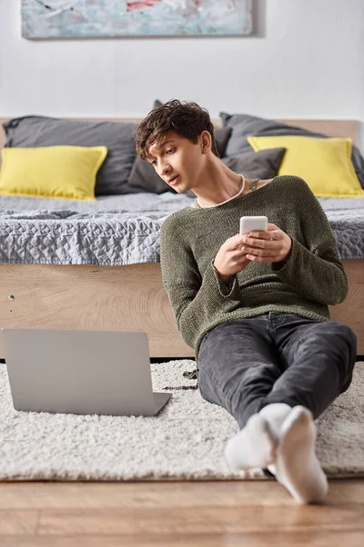Transgender-Bloggerin nutzt Smartphone und schaut auf Laptop, während sie auf Teppich sitzt, soziale Medien — Stockfoto