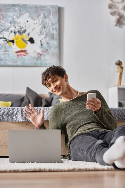 Curly transgender blogger holding smartphone and waving hand at laptop while sitting on carpet — Stock Photo