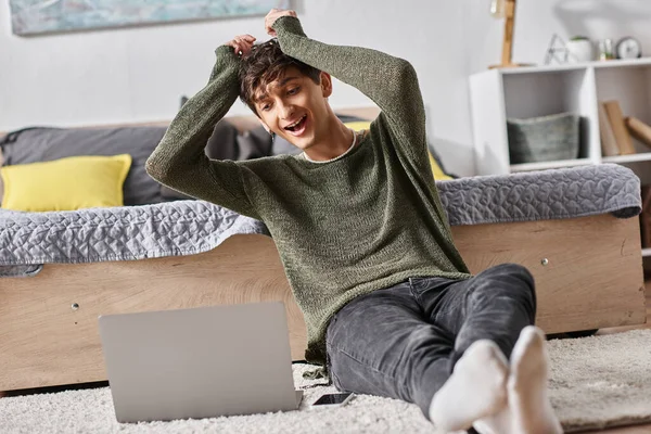 Sorprendido blogger transgénero tirando del pelo rizado y mirando a la computadora portátil mientras está sentado en la alfombra - foto de stock