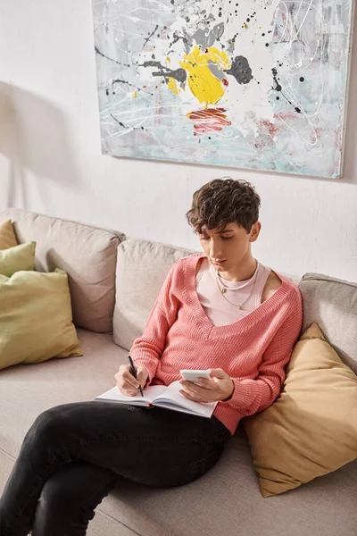 Трансгендерный блогер в розовом свитере держит смартфон и пишет в блокноте, сидит на диване — стоковое фото