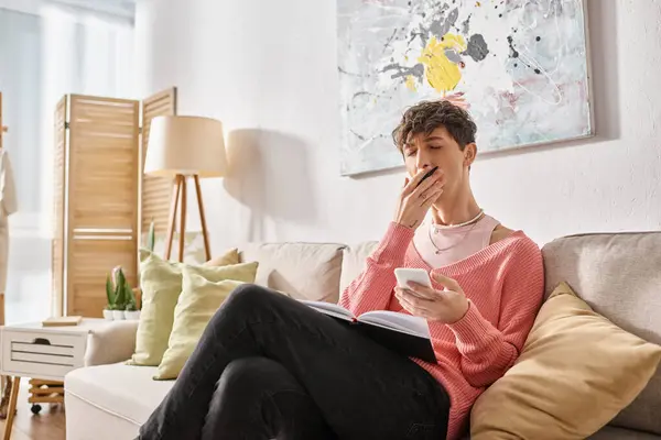 Tired transgender blogger in pink sweater holding smartphone and yawning while sitting on sofa — Stock Photo