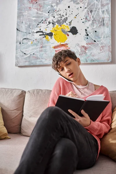 Трансгендерний блогер в рожевому светрі говорить на смартфоні і пише в блокноті, сидячи на дивані — стокове фото