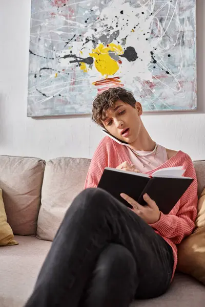 Transgender blogger in pink sweater talking on smartphone and writing in notebook, sitting on sofa — Stock Photo