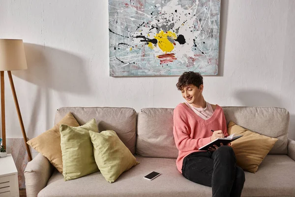 Felice transgender blogger in maglione rosa scrivere su notebook e guardare smartphone sul divano — Foto stock