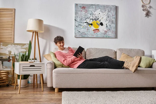 Happy transgender person in pink sweater writing in notebook and sitting on sofa next to smartphone — Stock Photo