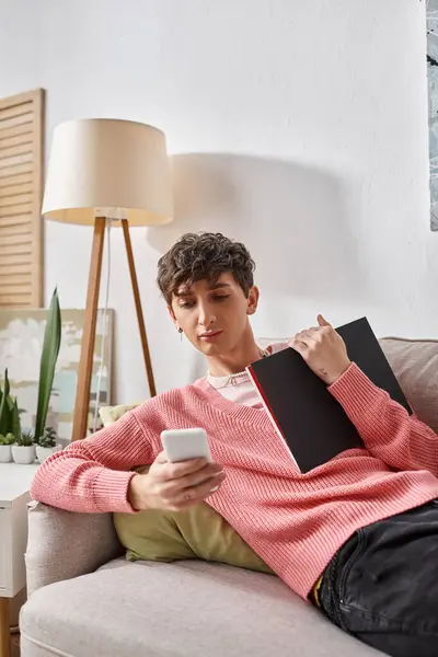 Encaracolado transgênero pessoa em rosa suéter segurando notebook, usando smartphone e sentado no sofá — Fotografia de Stock