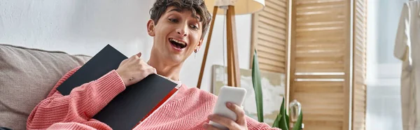 Amazed transgender blogger in pink sweater holding notebook and using smartphone, banner — Stock Photo