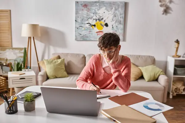 Joven transgénero freelancer en suéter rosa escritura en portátil cerca de la computadora portátil en el escritorio, trabajo remoto - foto de stock