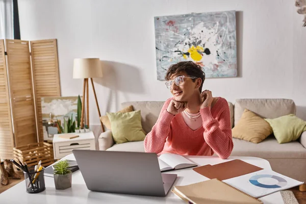 Felice transgender freelancer in maglione rosa guardando lontano vicino laptop sulla scrivania, lavoro a distanza — Foto stock