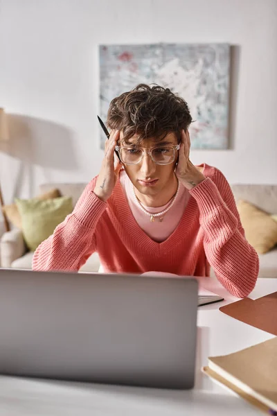 Young transgender freelancer in pink sweater and eyeglasses looking at laptop on desk, remote work — Stock Photo