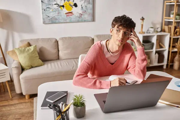 Pensive transgender freelancer in pink sweater and eyeglasses looking away while working remotely — Stock Photo