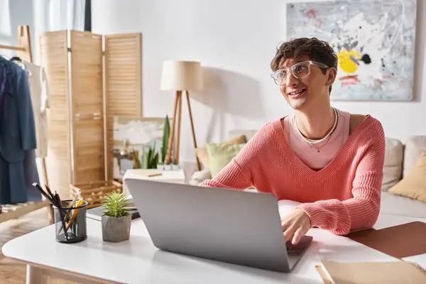 Happy androgynous freelancer in pink sweater and eyeglasses typing on laptop while working remotely — Stock Photo