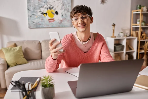 Happy androgynous freelancer in pink sweater and eyeglasses using smartphone near laptop on desk — Stock Photo
