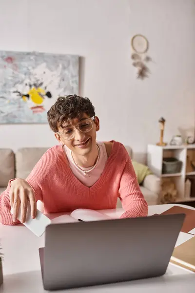Freelancer andrógino feliz em suéter rosa e óculos segurando smartphone perto de laptop na mesa — Fotografia de Stock