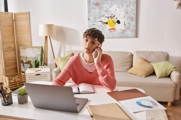 Curly androgynous freelancer in pink sweater and eyeglasses talking on smartphone near laptop — Stock Photo