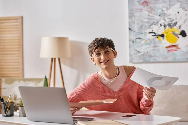 Счастливый андрогинный блогер в розовом свитере и очках работает дистанционно рядом с устройствами на столе — стоковое фото