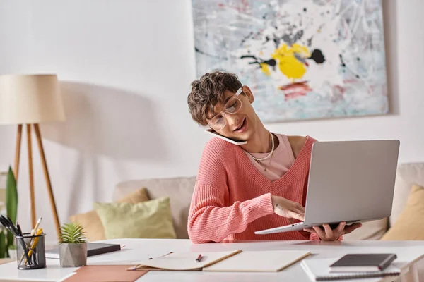 Allegro freelance androgino in maglione rosa e occhiali utilizzando laptop e smartphone a casa — Foto stock