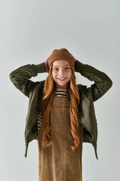Chica positiva con el pelo largo con sombrero de invierno y de pie en vestido con chaqueta sobre fondo gris - foto de stock