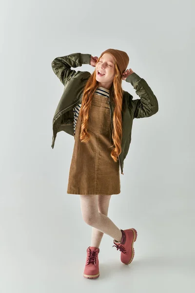 Chica alegre con el pelo largo con sombrero de invierno y de pie en vestido con chaqueta sobre fondo gris - foto de stock