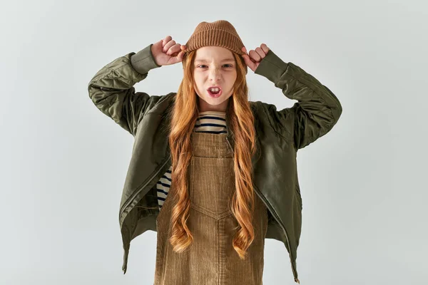 Chica emocional con el pelo largo con sombrero de invierno y de pie en vestido con chaqueta sobre fondo gris - foto de stock