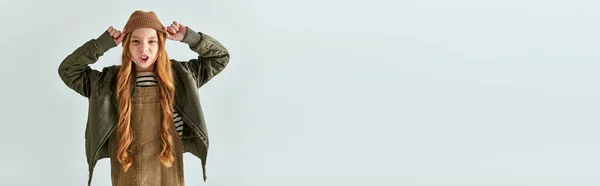 Fille émotionnelle avec les cheveux longs en robe avec veste chaude portant chapeau d'hiver sur fond gris, bannière — Photo de stock