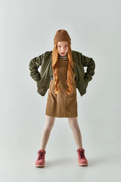 Chica enojada con el pelo largo y sombrero en la cabeza de pie en vestido con chaqueta de invierno sobre fondo gris — Stock Photo